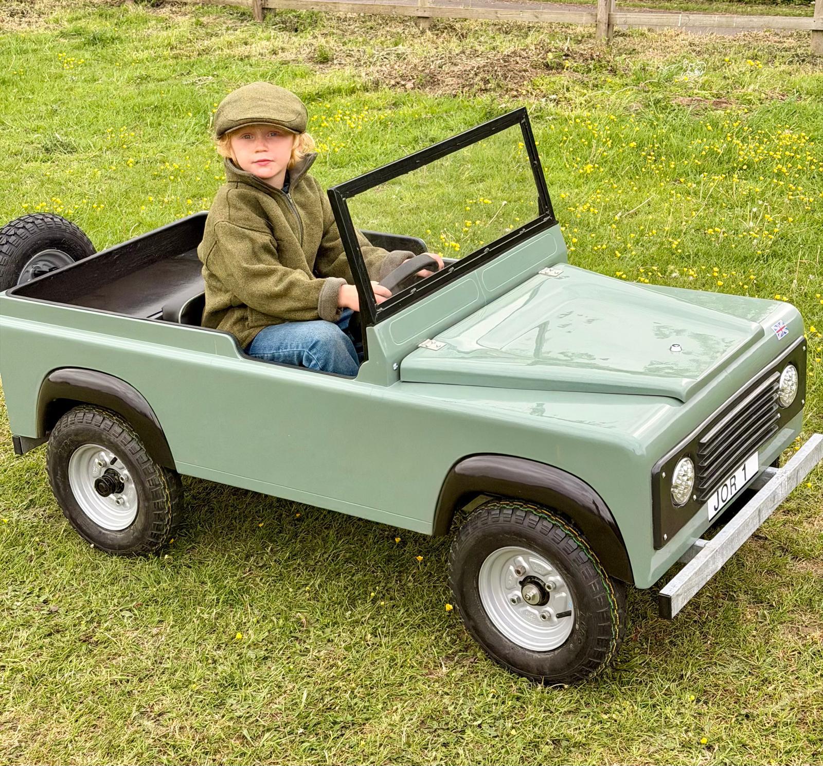 Child seated in a pastel green mini Land Rover, ready to drive